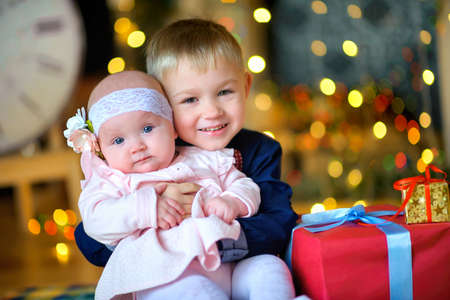 brother hugs his younger sister, sit on the floor near Christmas presents and gently smile, waiting for the holidaysの写真素材