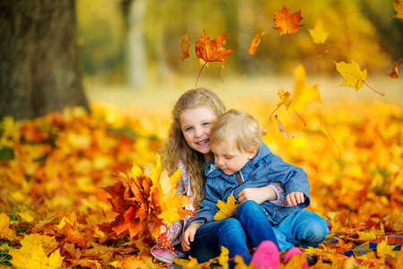 happy sister hugging her younger brother sitting in the park on fallen autumn foliageの写真素材