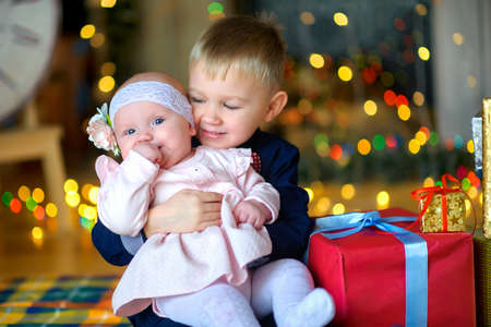 brother hugs his younger sister, sit on the floor near Christmas gifts, in the background a bright lightの写真素材