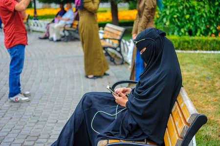 a modern Muslim woman in traditional hijab, sitting on a bench and playing on a smartphoneのeditorial素材
