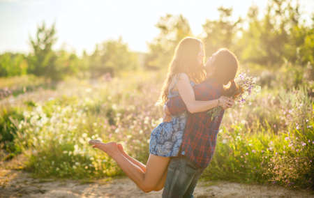 young happy couple is walking in the field, the guy is holding the girl in his arms in the rays of sunlightの写真素材