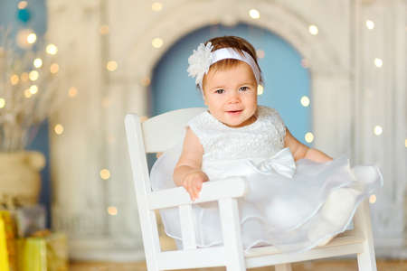 happy little girl in a dress, sits on a chair and smiles against a festive lightsの写真素材