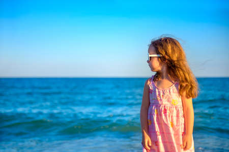 little beautiful girl in a dress standing on the beach looking away and dreaming of somethingの写真素材