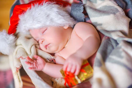 beautiful baby in Santa Claus hat lying asleep in basket, hugging a small box with a gift