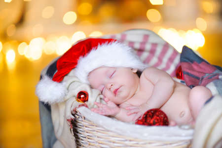 beautiful baby in a Santa Claus hat sipping lying in a basket, against a background of bright festive garlandsの写真素材