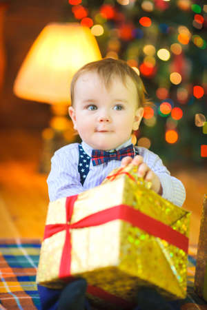 funny baby sitting on the floor with a big gift in hand, on the background of the festive Christmas treeの写真素材