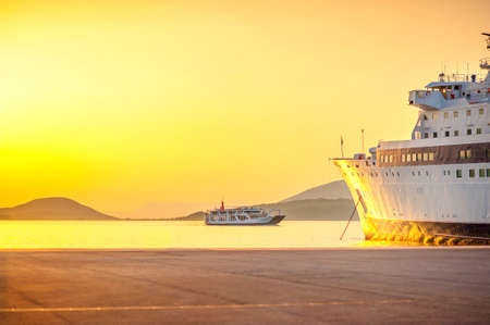 A large cargo ship standing in the port waiting for loading, against the background of a ferry sailing to the port, on a beautiful sunsetの写真素材
