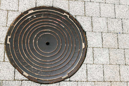 sewage dark sewer cover on the sidewalk between the blocks