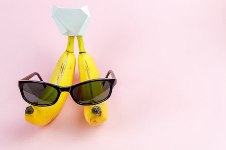 two torn bananas with a hat and sunglasses on a pink background with space for textの写真素材