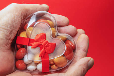 Colorful candy tablets in a heart-shaped plastic box with a ribbon on a red background held in the handsの写真素材
