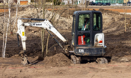 in early spring, a small bobcat excavator with rubber tracks in the park prepares the ground for the...