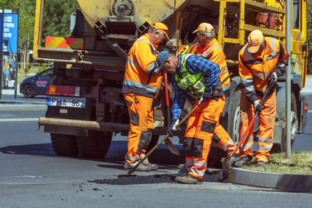 Road workers use shovels and rakes to repair asphalt on a street on a sunny summer day. Panevezys. Lithuania. 02. 06. 2023.のeditorial素材