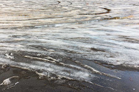 Spring brings melting snow and ice creating a beautiful scene on a black asphalt lot under a warm...