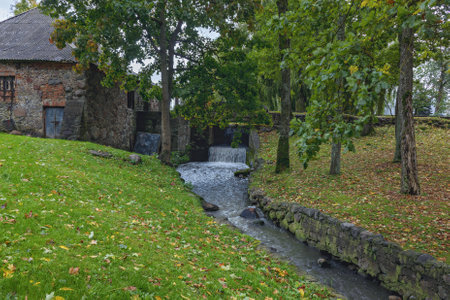 A small waterfall in the park flows over rocks into the river below. In the background stands a stone building with green trees under an overcast sky.の写真素材