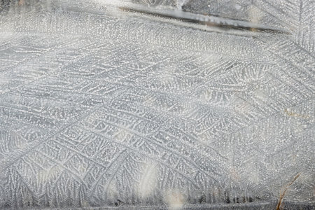 The frost creates delicate, lace-like patterns on the surface of the puddle, showing the artistry of...