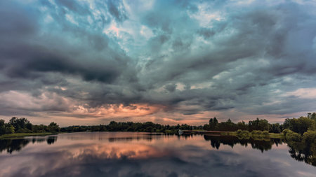 In the morning sky over a calm lake, golden hues blend with deep blues and grays. The surrounding trees frame the view, enhancing the tranquil atmosphere.の写真素材