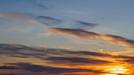 The sun sets on the horizon, casting warm hues of orange and yellow across the sky. Wispy clouds float gently, enhancing the beauty of the serene evening atmosphere.の写真素材