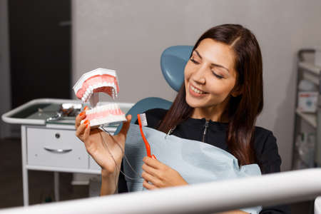 A young beautiful girl sits and smiles in a medical chair at the dentist's appointment and holds a mock jaw and a toothbrush in her hands.の写真素材