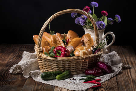 Wicker basket with vegetables and pastries, tablecloth and vase with flowers on an old wooden deskの写真素材