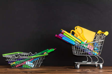The concept of going back to school. Space for text. Office supplies in a shopping basket on a wooden table near the blackboardの写真素材