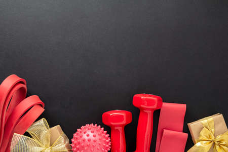 Fitness concept merry Christmas and Happy New Year. Red dumbbells, a fitness elastic band, a massage ball and golden gifts with ribbons on black. Flat lay, top view, copy spaceの写真素材