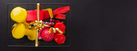 A festive set of tools for sports. Red and yellow dumbbells, fitness bands, gloves and a massage ball lie in a metal basket on a black background. Top view, flat lay, copy space, space for textの写真素材