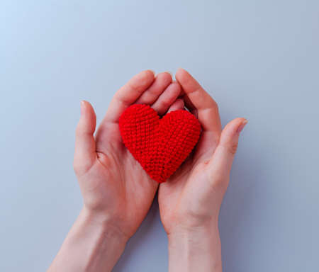 Valentines day background. Womens hands hold a red knitted heart made of threads on a blue background. Close-up. The concept of a holiday and relationships. Flat lay, copy spaceの写真素材