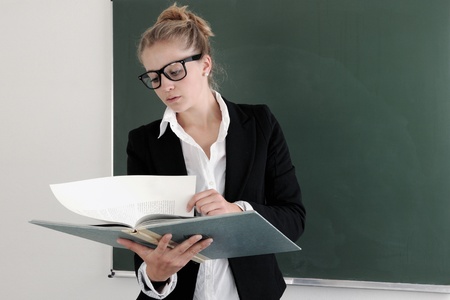 Portrait of young teacher with book and eyeglasses looking at camera by the blackboard の写真素材