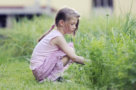 Adorable little girl in colorful clothes sits on grass in front of old style houseの写真素材
