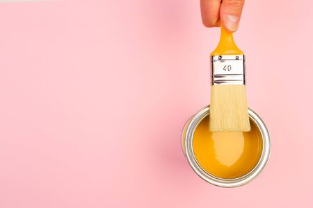 hand holding Brush  on open can of yellow  paint on pastel pink background. Renovation conceptの写真素材