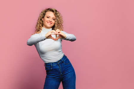 beautiful young woman   with curl hair  posing  on pink   background  and making heart with handsの写真素材