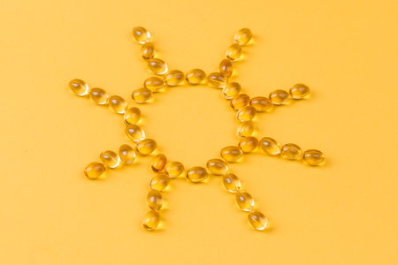 Yellow oil capsules with vitamin D  forming shape of sun on yellow background.