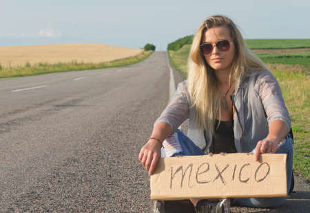 Serious blonde holding sign while hitchhiking on the road in summertime.の写真素材