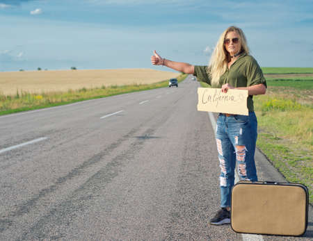 Blonde holding sign while hitchhiking on the road in summertime.の写真素材