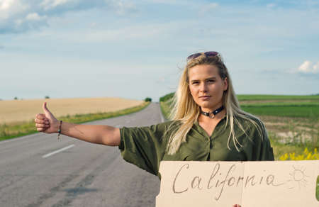 Blonde holding sign while hitchhiking on the road in summertime.の写真素材