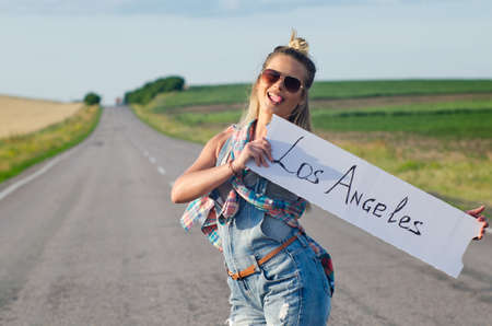 Blonde holding sign while hitchhiking on the road in summertime.の写真素材