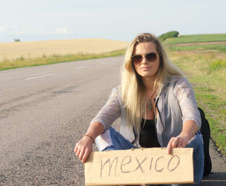 Blonde holding sign while hitchhiking on the road in summertime.の写真素材