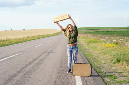 Blonde holding sign while hitchhiking on the road in summertime.の写真素材