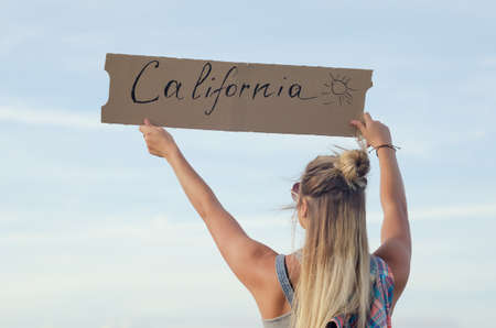 Blonde holding sign while hitchhiking on the road in summertime.の写真素材