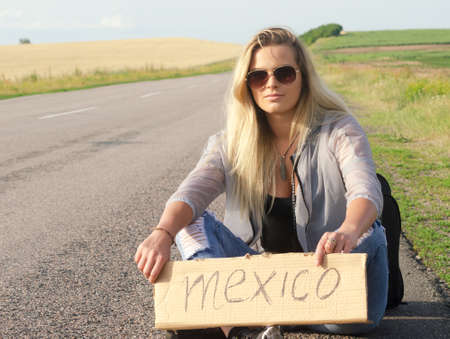 Blonde holding sign while hitchhiking on the road in summertime.の写真素材