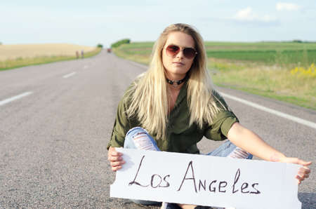 Blonde holding sign while hitchhiking on the road in summertime.の写真素材