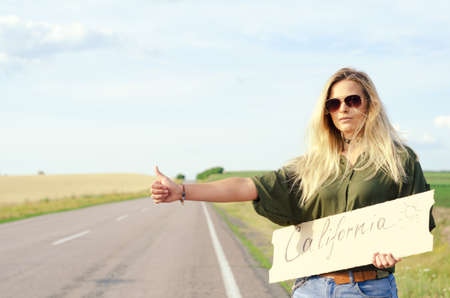 Blonde holding sign while hitchhiking on the road in summertime.の写真素材