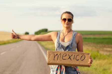 Blonde holding sign while hitchhiking on the road in summertime.の写真素材