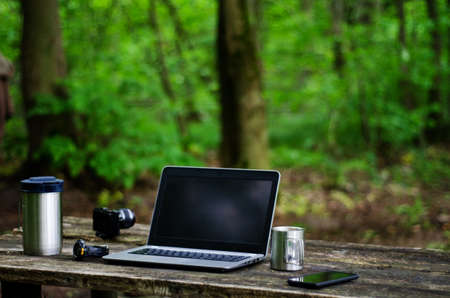 Close-up photo of a laptop, camera, coffee and smartphone on a wooden table outdoors.