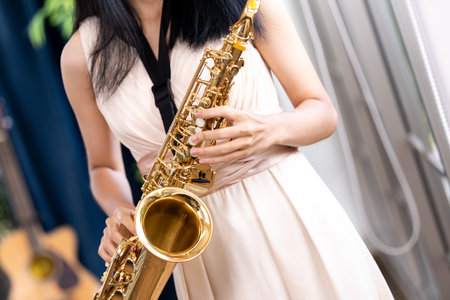 Young woman saxophonist with an instrument stands by the window. Cozy home interior. Concept of hobbies, inner world, development of talents, music school.の写真素材