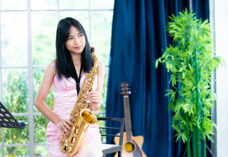 Young woman saxophonist with an instrument stands by the window. Cozy home interior. Concept of hobbies, inner world, development of talents, music school.の写真素材