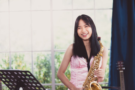 Young woman saxophonist with an instrument stands by the window. Cozy home interior. Concept of hobbies, inner world, development of talents, music school.の写真素材