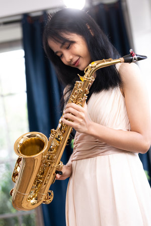 Young woman saxophonist with an instrument stands by the window. Cozy home interior. Concept of hobbies, inner world, development of talents, music school.の写真素材