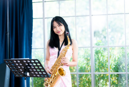 Young woman saxophonist with an instrument stands by the window. Cozy home interior. Concept of hobbies, inner world, development of talents, music school.の写真素材