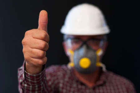 Worker shows thumbs up, with hard hat, safety glasses, protective mask, selective focus and black background.の写真素材